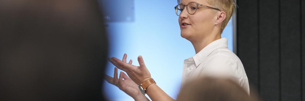 Person in a white shirt speaking and gesturing to an audience in front of a blue presentation screen.