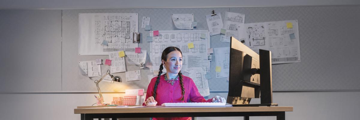 A person in a bright pink jumper sits at a desk using a computer, with plans pinned to the wall behind them.