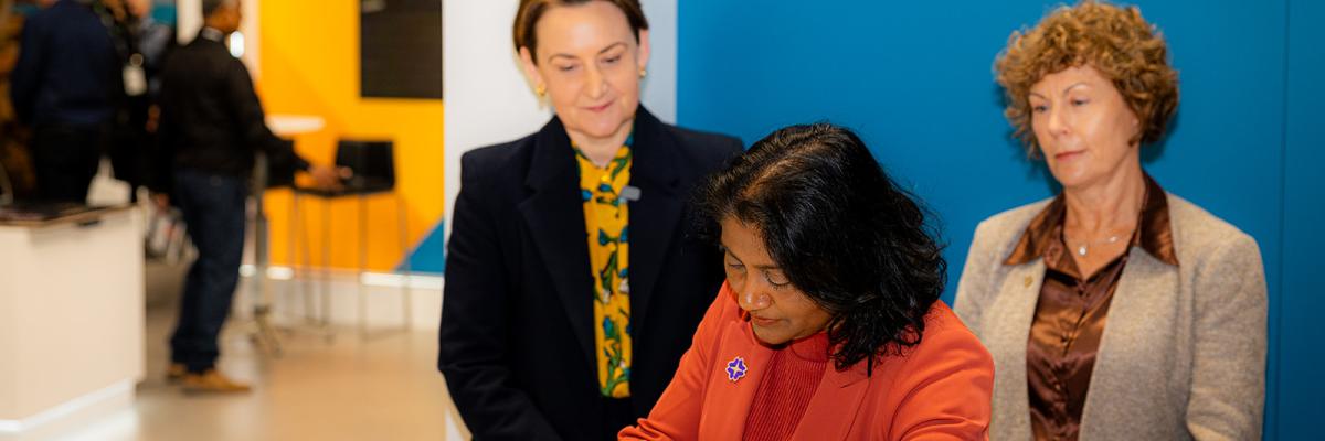 A person in an orange blazer signs a document at a white counter with two attendees standing nearby. 