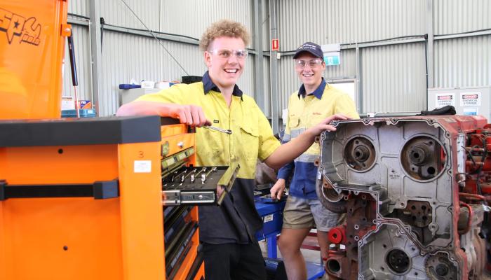 Two tradies smiling at the camera with car motor in foreground