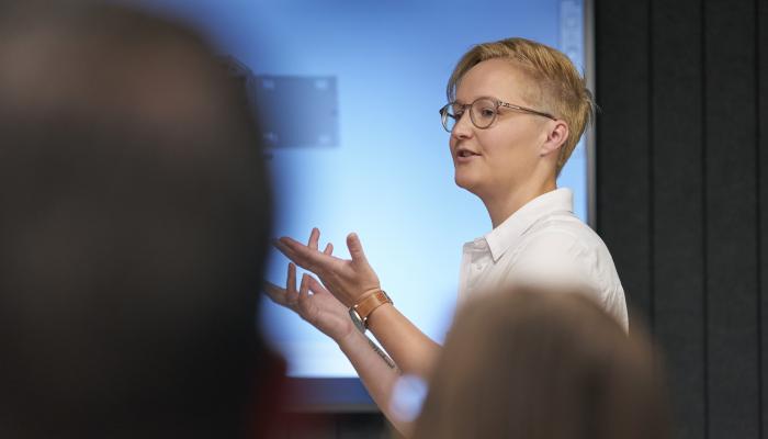 Person in a white shirt speaking and gesturing to an audience in front of a blue presentation screen.