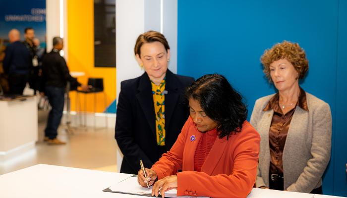 A person in an orange blazer signs a document at a white counter with two attendees standing nearby. 