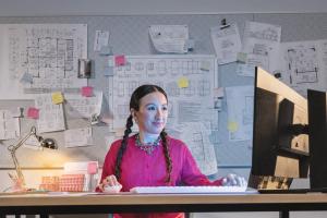 A person in a bright pink jumper sits at a desk using a computer, with plans pinned to the wall behind them.
